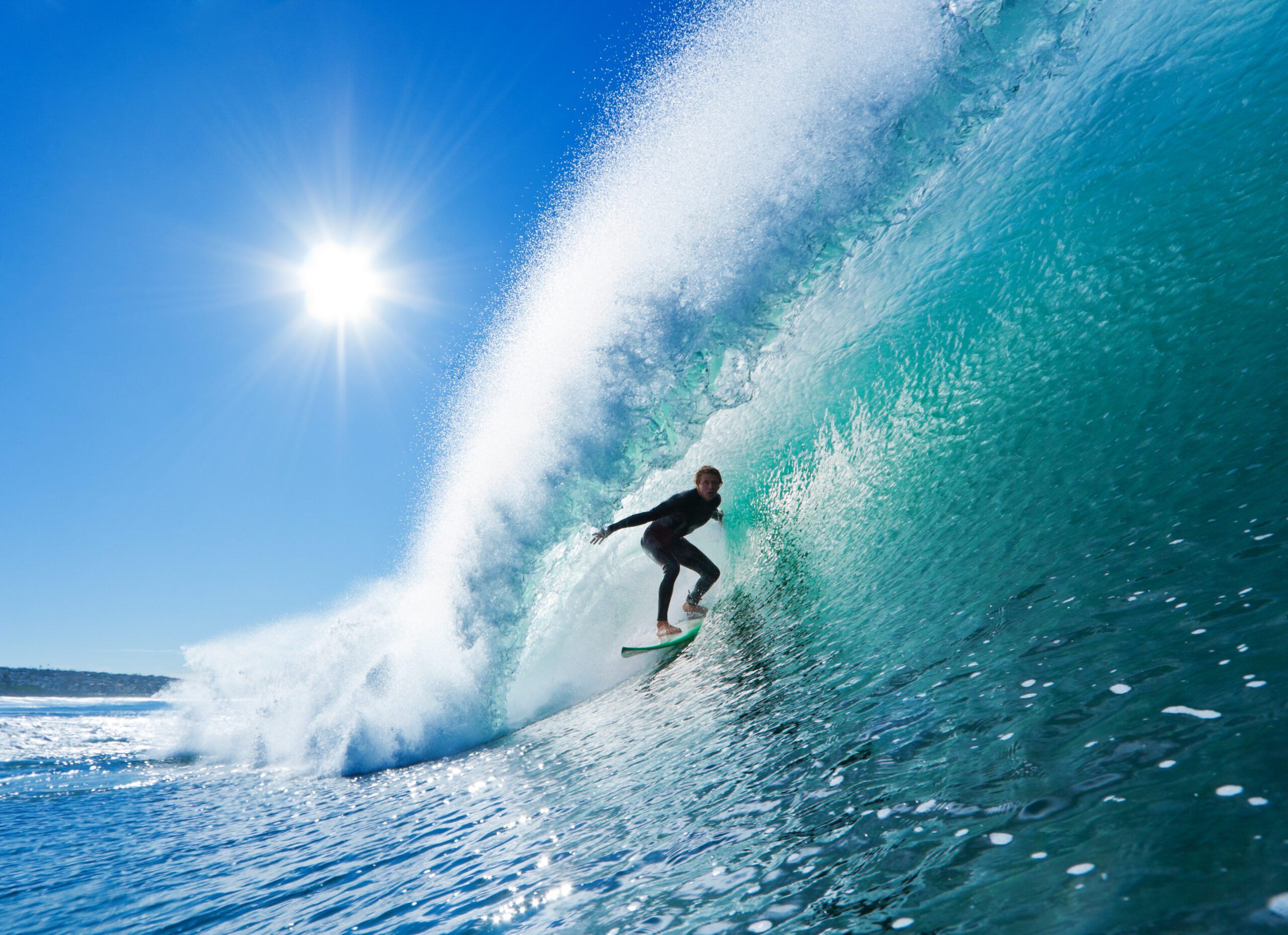 person surfing a large wave with the sun behind and a blue sky